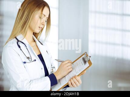 Young woman-doctor is making some notes using a clipboard, while standing in her cabinet in a clinic. Portrait of beautiful female physician with a Stock Photo