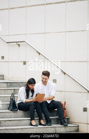 Young smiling entrepreneurs sitting on steps outdoors and discussing article on tablet computer Stock Photo