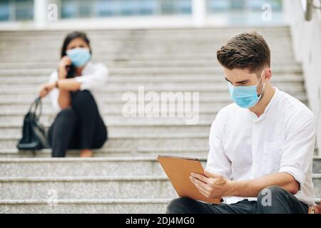 Serious young man in medical mask sitting on steps and reading article on tablet computer, his colleagues sitting in background Stock Photo