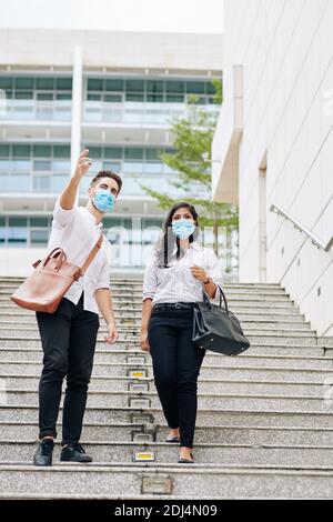 Young businessman in medical mask showing headquarters complex to new employee Stock Photo