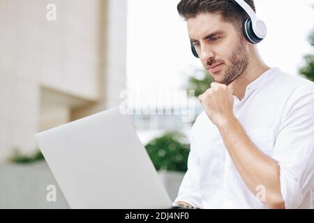 Pensive handsome young software developer in headphones rubbing chin and looking at laptop screen Stock Photo