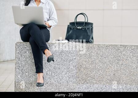Cropped image of young female entrepreneur sitting outdoors next to her bag and worrking on laptop Stock Photo