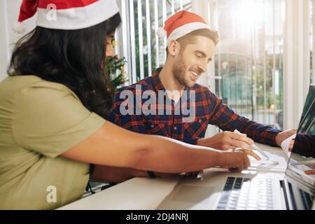 Happy young businessman in Santa Claus hat discussing sales report and business statistics with colleague at meeting Stock Photo