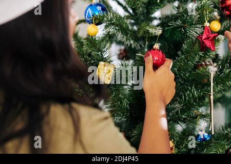 Young woman in Santa Claus hat hanging red bauble on Christmas tree Stock Photo