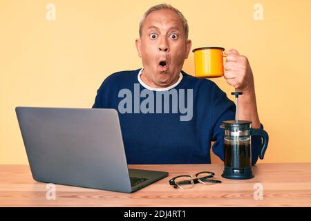 Senior handsome man with gray hair working at the office drinking a cup of coffee scared and amazed with open mouth for surprise, disbelief face Stock Photo