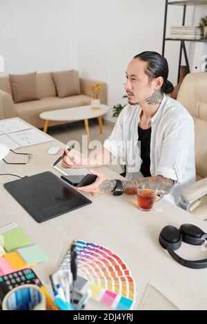 Experienced designer with smartphone in hands working on graphic tablet at his home office Stock Photo