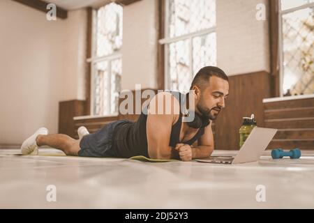 Laying side of a young athletic man in a black sports outfit, laying on the floor on a pad and watches video instructions using laptop. Video lessons Stock Photo