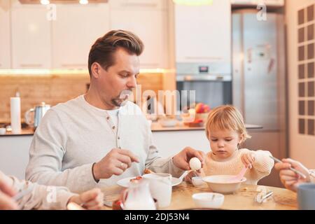 Portrait of modern family enjoying breakfast together in kitchen , focus on father helping cute baby girl, copy space Stock Photo