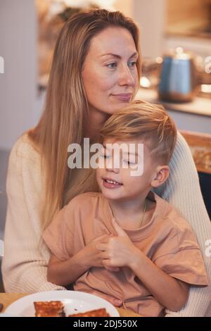 Vertical portrait of happy mother holding cute son while enjoying breakfast with family in cozy kitchen interior Stock Photo