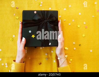 New Year or Christmas flat lay. Top view Xmas holiday celebration decorative black gift box with black bow, bright sparkles on the yellow background w Stock Photo
