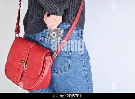 A girl in a mimes fit jeans, a sweater and with a red bag on the edge pulls out a retro audio cassette from her pocket on a white background. Side vie Stock Photo
