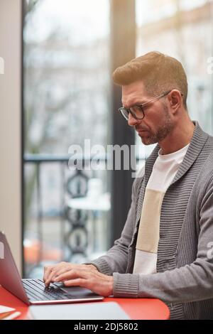 Vertical side view portrait of handsome mature man using laptop while working at home office against window Stock Photo