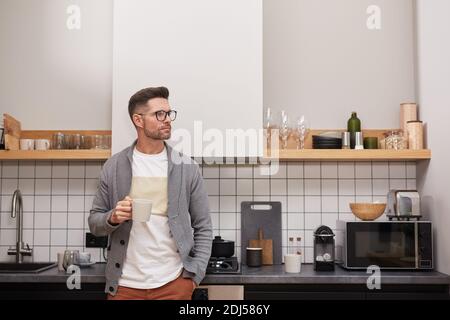 Waist up portrait of elegant adult man holding coffee mug and looking away pensively while relaxing standing in kitchen interior, copy space Stock Photo