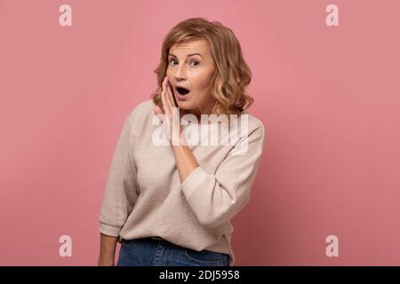 Senior woman with hand on mouth telling secret rumor, whispering malicious talk conversation. studio shot on pink wall. Stock Photo