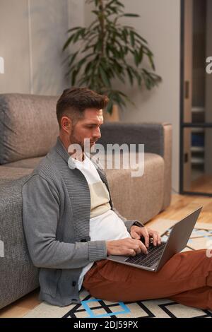Vertical side view portrait of handsome mature man working with laptop while sitting on floor at home in modern interior Stock Photo