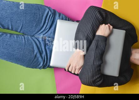 A girl in jeans and a sweater lies on a colored paper background and hugs a laptop. Top view. Stock Photo