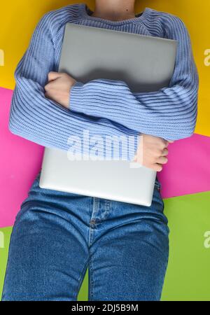 A girl in jeans and a sweater lies on a colored paper background and hugs a laptop. Top view. Stock Photo