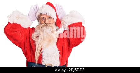 Old senior man with grey hair and long beard wearing traditional santa claus costume doing bunny ears gesture with hands palms looking cynical and ske Stock Photo