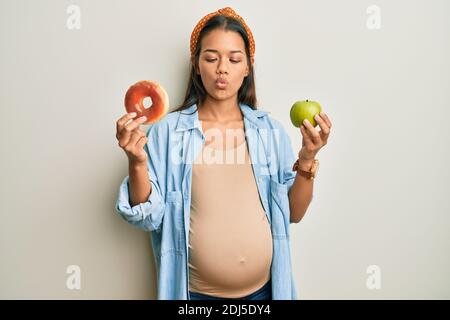 Beautiful hispanic woman expecting a baby, holding food making fish face with mouth and squinting eyes, crazy and comical. Stock Photo