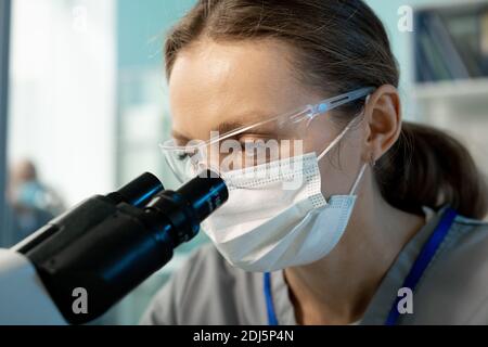 Young contemporary female scientist in protective mask and eyewear looking at chemical or biological samples in microscope in laboratory Stock Photo