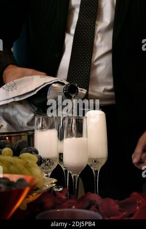 Man pours sparkling wine into glasses on the table in the middle of rose petals and fruit Stock Photo