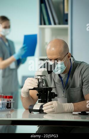 Mature bald male scientist in protective mask and gloves studying chemical samples in microscope against female assistant working with documents Stock Photo