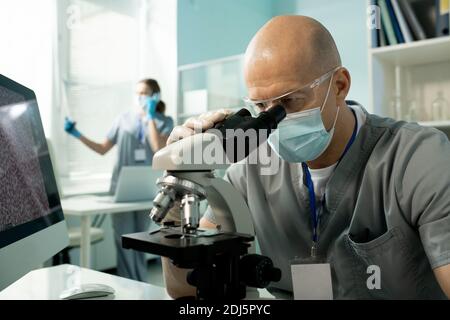 Mature bald male clinician in protective mask and gloves studying new vaccine in microscope against female assistant talking on the phone Stock Photo