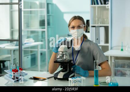 Young female scientist or clinician in protective workwear looking at you while using in microscope during scientific research in laboratory Stock Photo