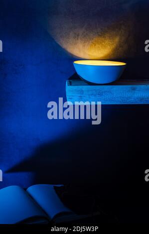 a high contrast image of a bamboo bowl glowing bright yellow on a wooden shelf above a desk. On the desk lays an open notebook with blank pages Stock Photo
