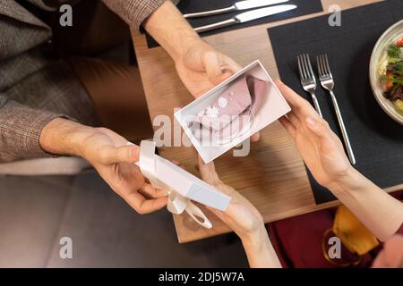 Hands of young man holding open giftbox with handmade protective mask decorated with beads while giving it to his girlfriend in restaurant Stock Photo