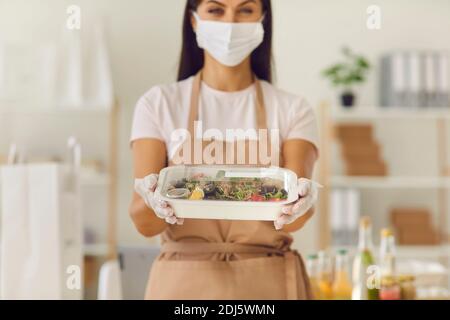 Woman in a mask gives the client a ready fresh healthy lunch in a plastic container. Stock Photo