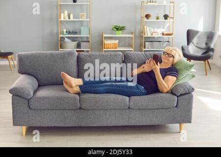 Positive senior woman lying on sofa, relaxing and chatting or making order online on smartphone Stock Photo