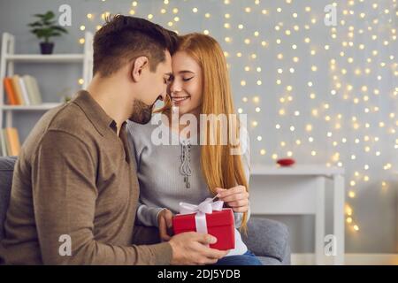 Young loving happy couple woman and man sitting together on sofa and opening holiday present box Stock Photo
