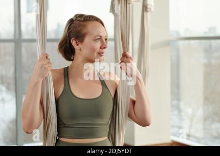 Young happy sportswoman in grey tracksuit holding by fly yoga hammock in gym while exercising in front of camera and looking through window Stock Photo