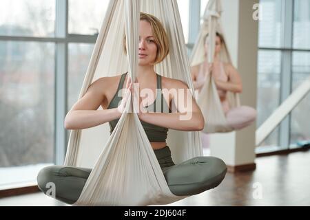 Young fit woman in tracksuit sitting in pose of lotus with her legs crossed and hands put together by chest while exercising in gym Stock Photo