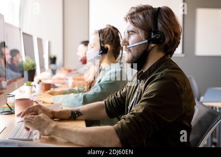 Young serious bearded male operator with protective mask under chin consulting clients while sitting in front of colleagues in headsets Stock Photo