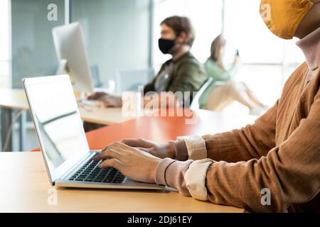 Hands of businesswoman in protective mask and casualwear pressing laptop keypad buttons while sitting by desk against colleagues Stock Photo