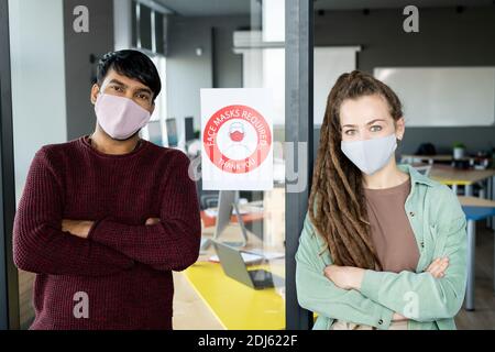 Two young office workers in masks and casualwear crossing arms on chest while leaning against announcement with requirement in open space Stock Photo
