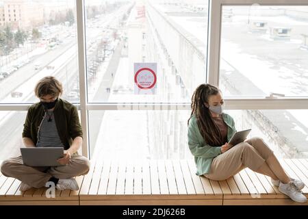 Two young office workers in casualwear sitting against large window with announcement about keeping distance and using mobile gadgets Stock Photo