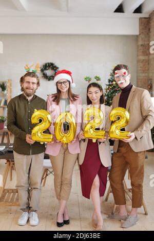 Several happy young white collar workers in xmas accessories holding inflatable numbers of new year while standing in front of camera Stock Photo