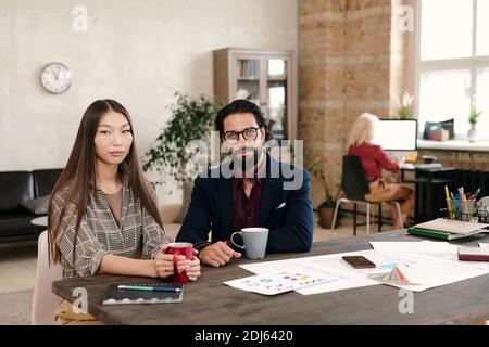 Young Asian female office worker and her bearded colleague having coffee at break while sitting by table with many papers against businesswoman Stock Photo