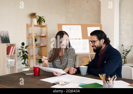 Young Asian female office worker pointing at sketch on paper and her happy bearded colleague looking at it while preparing presentation Stock Photo