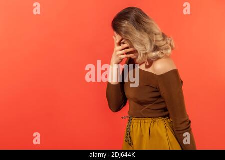 Side view portrait of unhappy young woman with blonde curly hair in casual clothes standing and covering with hand on head, feeling sorrow. Indoor stu Stock Photo