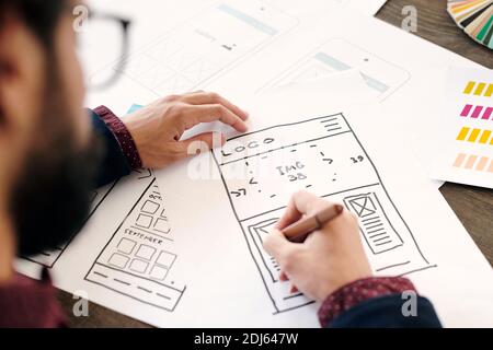 Hand of young contemporary developer of mobile application holding pencil over sketch on paper while working over one of new items Stock Photo