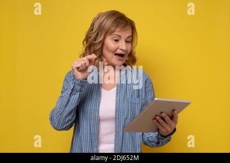 Mature attractive smiling woman using a tablet computer selecting goods in online shop on a yellow background Stock Photo