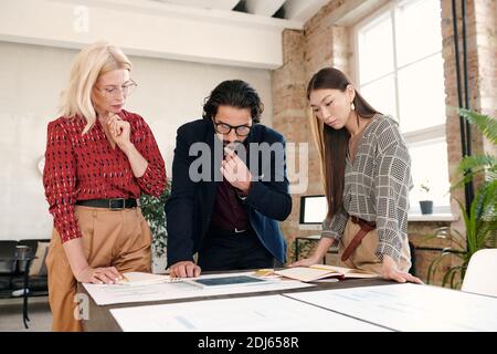 Three pensive intercultural mobile application developers standing by table and bending over papers with sketches while looking at tablet display Stock Photo