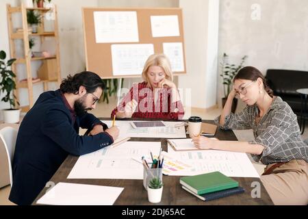 Three pensive colleagues sitting by table and brainstorming while working individually in office environment against board with sketches Stock Photo