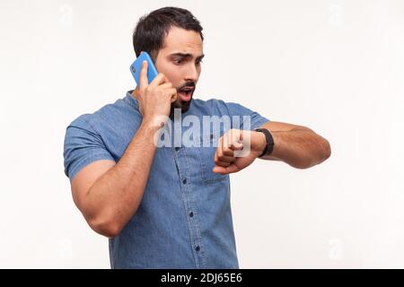 Impatient bearded business man talking phone and checking time looking at his smartwatch, shocked and confused with time, deadline, time to go. Indoor Stock Photo