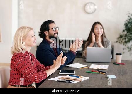 Three happy intercultural professionals clapping their hands while sitting by table and expressing satisfaction with presentation or speech Stock Photo