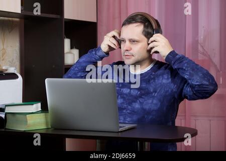 A man in home clothes works at a laptop. Puts headphones on his head. There are books on the table nearby. Stock Photo